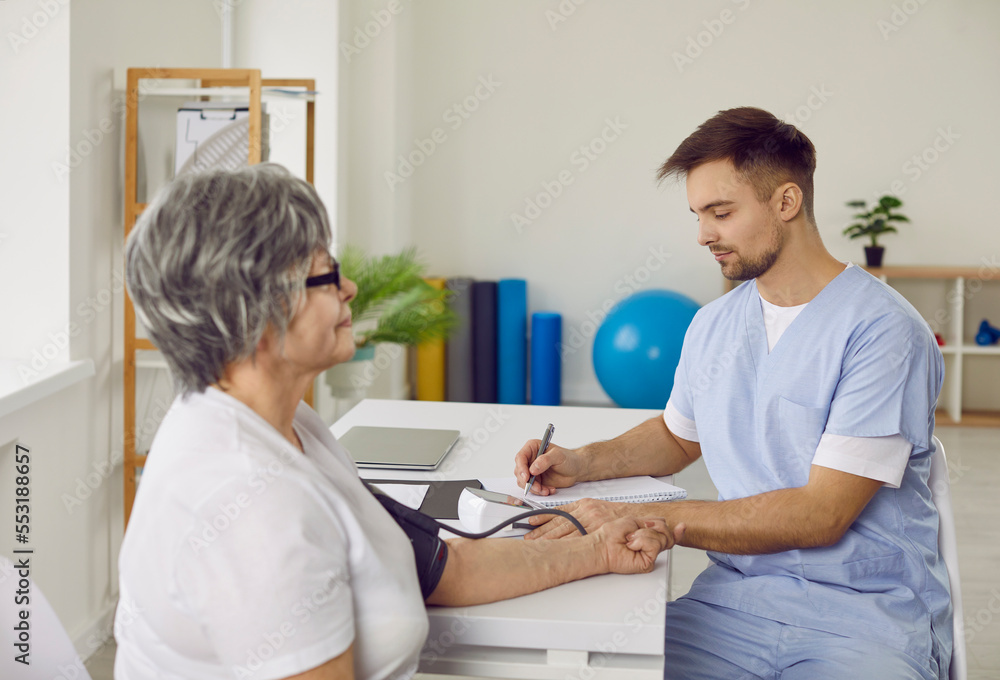 Fototapeta premium Young doctor measuring arterial blood pressure of senior patient. Elderly woman sitting at desk with tonometer cuff on her arm during visit to hospital. Health, medicine, disease prevention concept