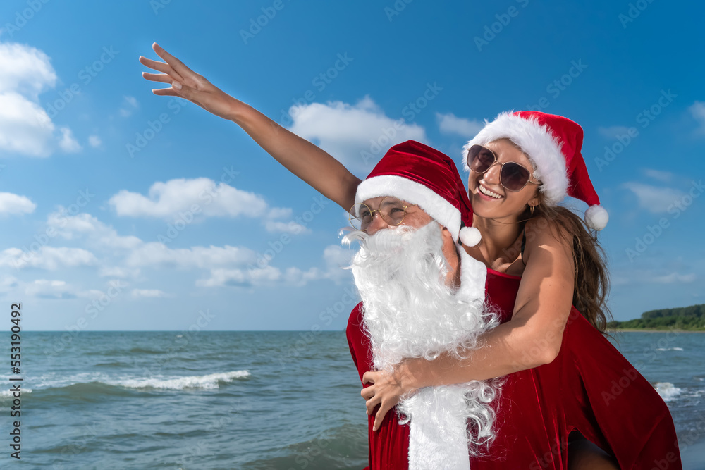 Obraz premium Santa Claus is holding on his back a young smiling woman in a Santa hat pointing her finger to the left on the seashore on a sunny day. Concept of celebrating Christmas holidays in warm countries.