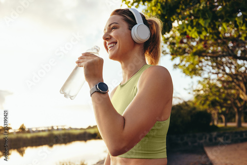 Fototapeta Female athlete taking a break outdoors, drinking water and listening to music wi