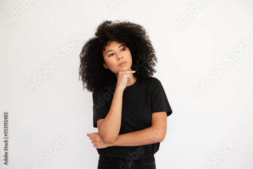 Pensive young woman looking away holding thumb on her chin. Hispanic female model with afro hairstyle and brown eyes in black T-shirt looking away touching chin with thumb. Thought, dream concept © KAMPUS