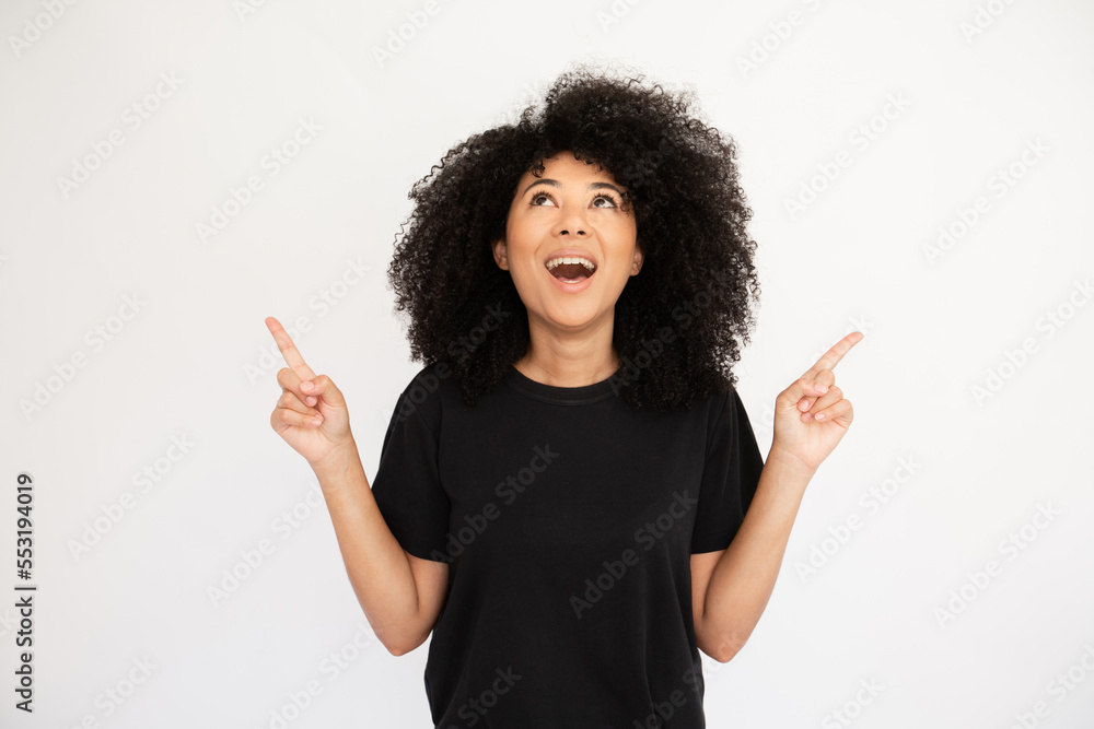 Amazed young woman pointing above at copyspace. Caucasian female model with afro hairstyle and brown eyes in black T-shirt opening mouth from surprise presenting advertisement. Advertising concept Amazed young woman pointing above at copyspace. Caucasian female model with afro hairstyle and brown eyes in black T-shirt opening mouth from surprise presenting advertisement. Advertising concept