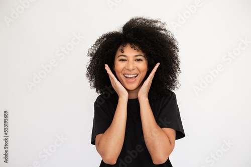 Amazed young woman throwing hands up. Caucasian female model with afro hairstyle and brown eyes in black T-shirt smiling and opening her mouth from surprise. Emotion, astonishment concept
