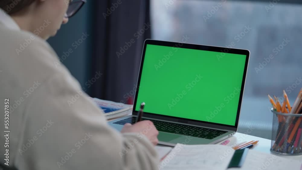 Woman Uses Laptop with Green Mock-up Screen. Girl Student Taking Notes on Notebook from Laptop at Online Lesson Woman Uses Laptop with Green Mock-up Screen. Girl Student Taking Notes on Notebook from Laptop at Online Lesson