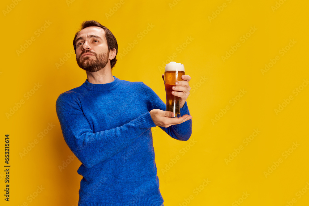 Portrait of emotive man in blue sweater posing, proudly holding glass with lager foamy beer isolated over yellow background Portrait of emotive man in blue sweater posing, proudly holding glass with lager foamy beer isolated over yellow background