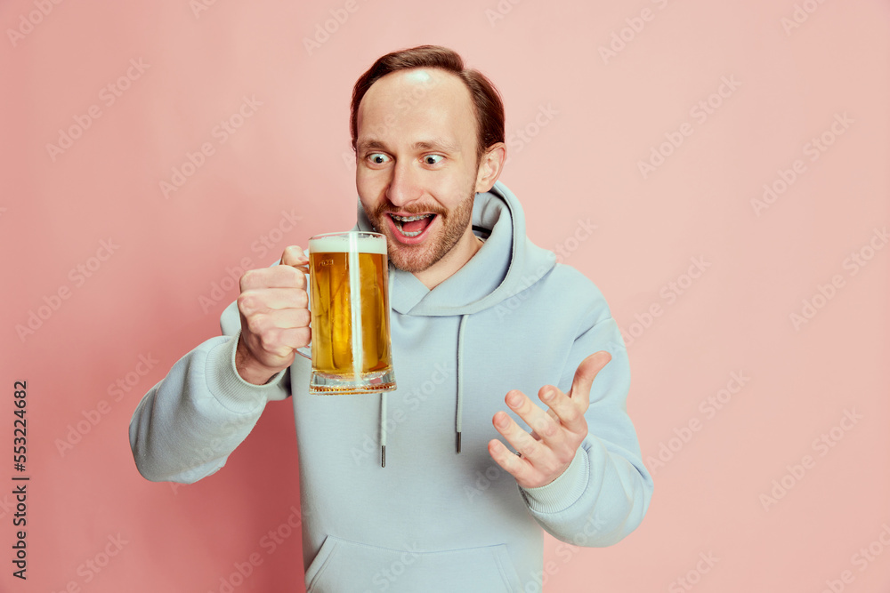 Portrait of young man in casual hoodie posing with lager foamy beer mug isolated over pink background. Looking with excitement Portrait of young man in casual hoodie posing with lager foamy beer mug isolated over pink background. Looking with excitement
