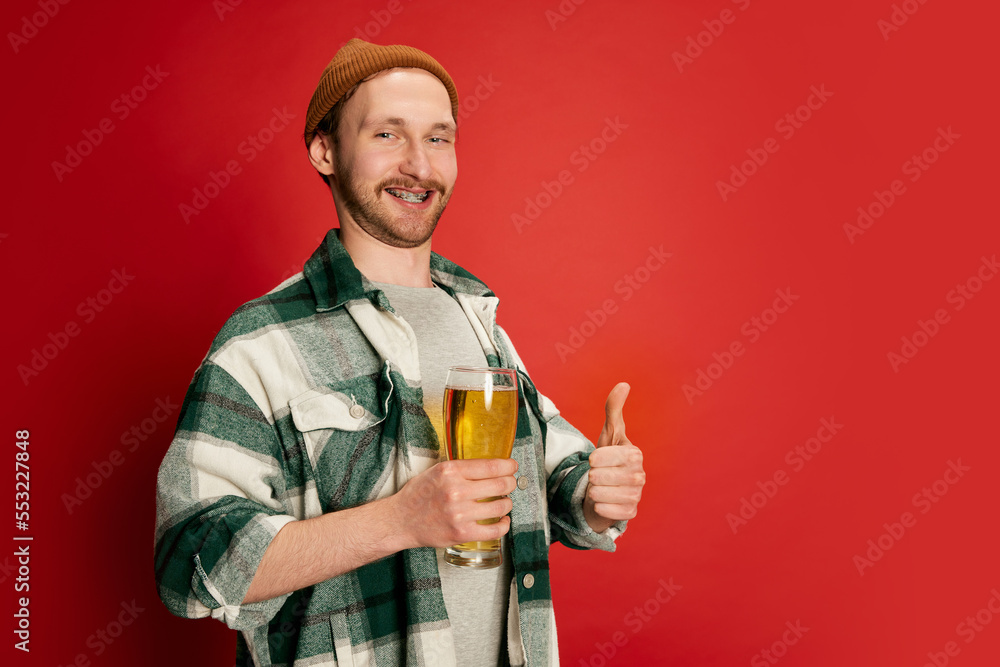 Portrait of young man in casual checkered shirt posing with beer glass isolated over red background. Looking happy. Perfect taste Portrait of young man in casual checkered shirt posing with beer glass isolated over red background. Looking happy. Perfect taste