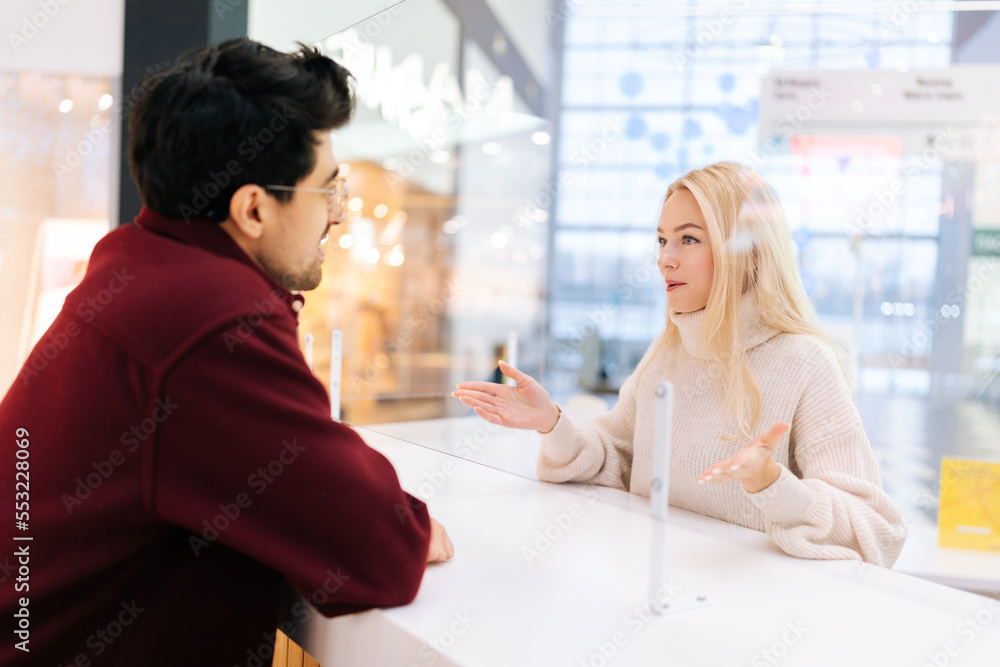 Rear view from shoulder of smiling man talking through glass partition with confused blonde woman standing at desk in bright hall of shopping mall. Concept of lifestyle social distancing and work. Rear view from shoulder of smiling man talking through glass partition with confused blonde woman standing at desk in bright hall of shopping mall. Concept of lifestyle social distancing and work.