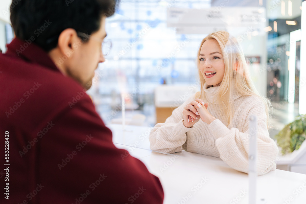 Back view from shoulder of serious man in glasses talking through glass partition with blonde woman standing at desk in bright hall of shopping mall. Concept of lifestyle social distancing and work.