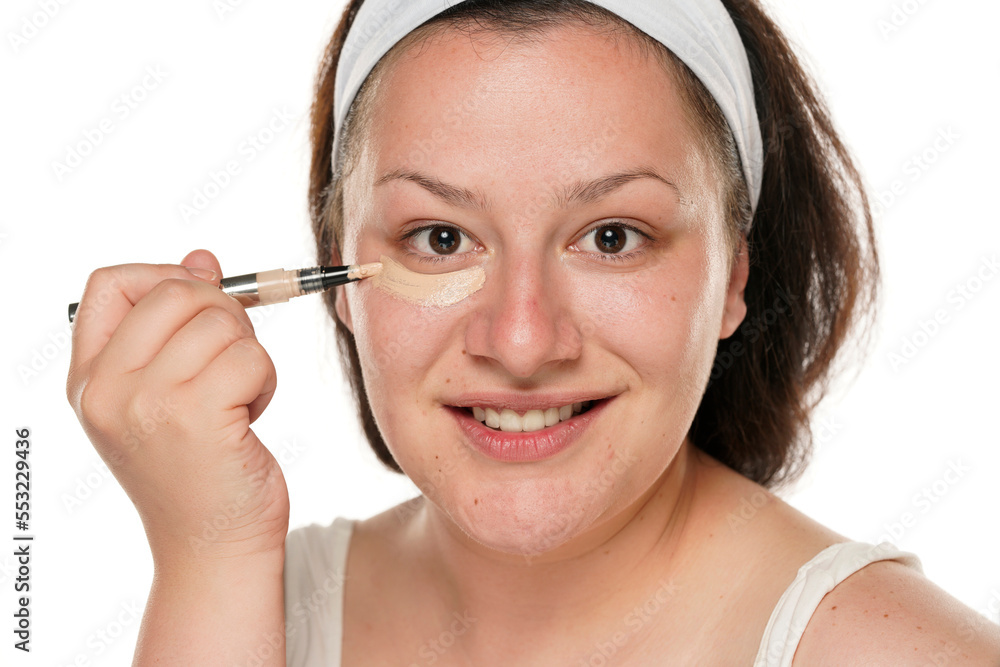 Portrait of a young smiling chubby woman applyes concealer under her eyes on white background. Portrait of a young smiling chubby woman applyes concealer under her eyes on white background.