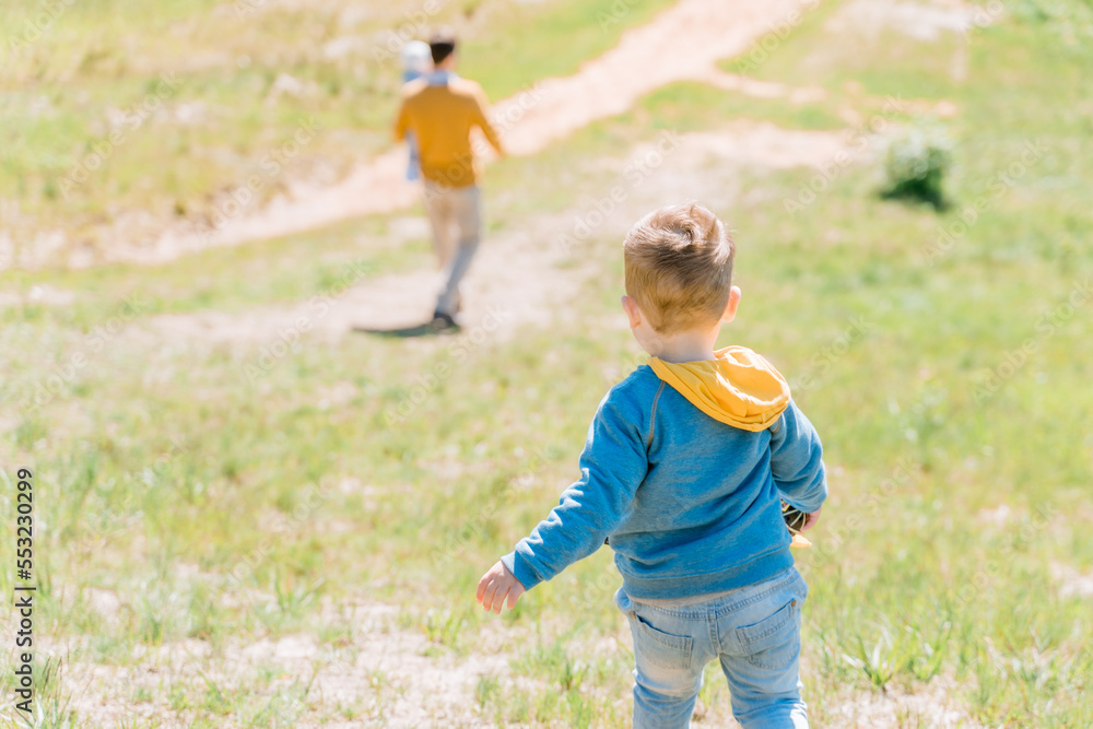 A boy of three years old follows his father carrying his youngest daughter in his arms along a green hilly lawn on a sunny day, view from the back A boy of three years old follows his father carrying his youngest daughter in his arms along a green hilly lawn on a sunny day, view from the back