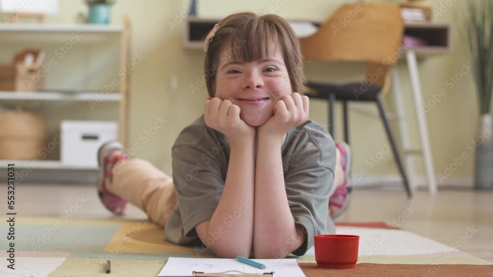 Portrait of pretty Caucasian girl with down syndrome smiling and showing funny faces at camera while lying on her stomach on rug in living room Portrait of pretty Caucasian girl with down syndrome smiling and showing funny faces at camera while lying on her stomach on rug in living room