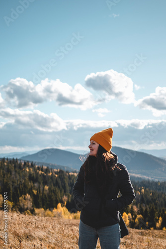 Young happy woman in orange beanie is standing in front of mountains landscape