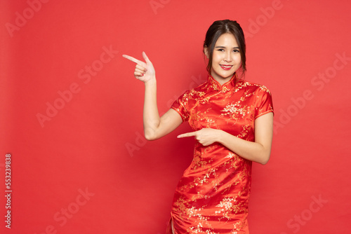Young Asian woman wearing red traditional cheongsam qipao dress smiling and pointing to empty copy space isolated on red background photo