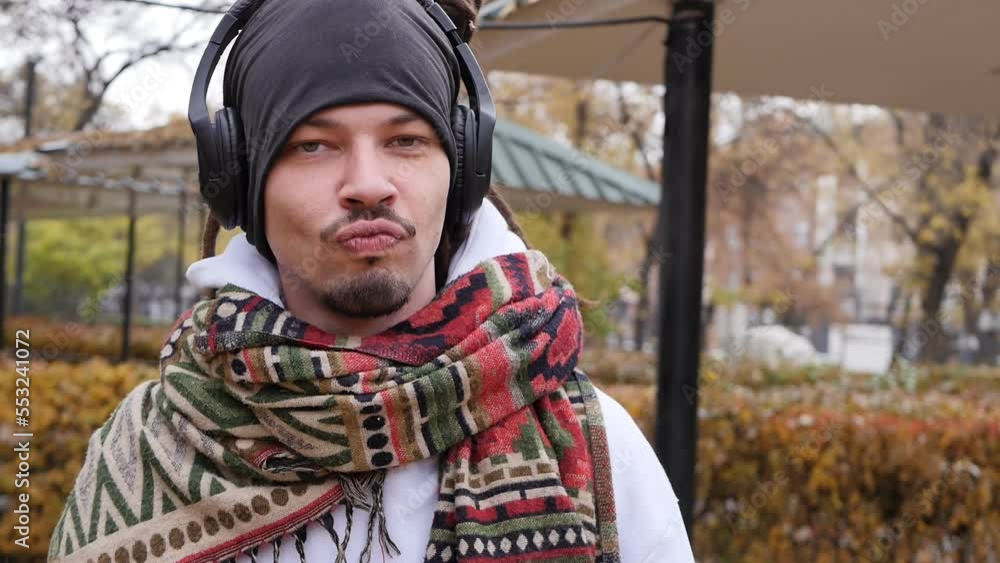 A handsome young man with dreadlocks and a scarf wound around his neck listens to music using headphones while having fun and moving his head to the beat of the melody outdoors A handsome young man with dreadlocks and a scarf wound around his neck listens to music using headphones while having fun and moving his head to the beat of the melody outdoors