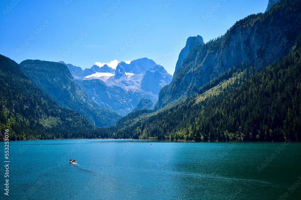 The stunnnig summer Alpine landscape with the mountain background and the lake, with boat roaming through the deep, perfectly clean blue waters 