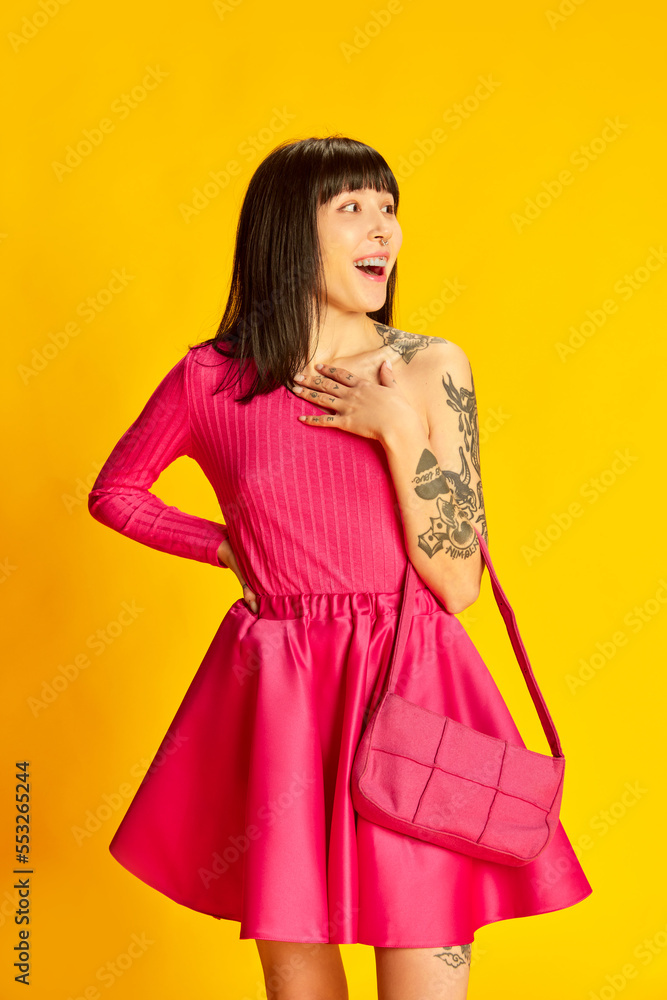 Portrait of young beautiful girl in bright pink dress posing, cheerfully smiling isolated on vivid yellow background. Concept of youth, beauty, fashion, lifestyle, emotions
