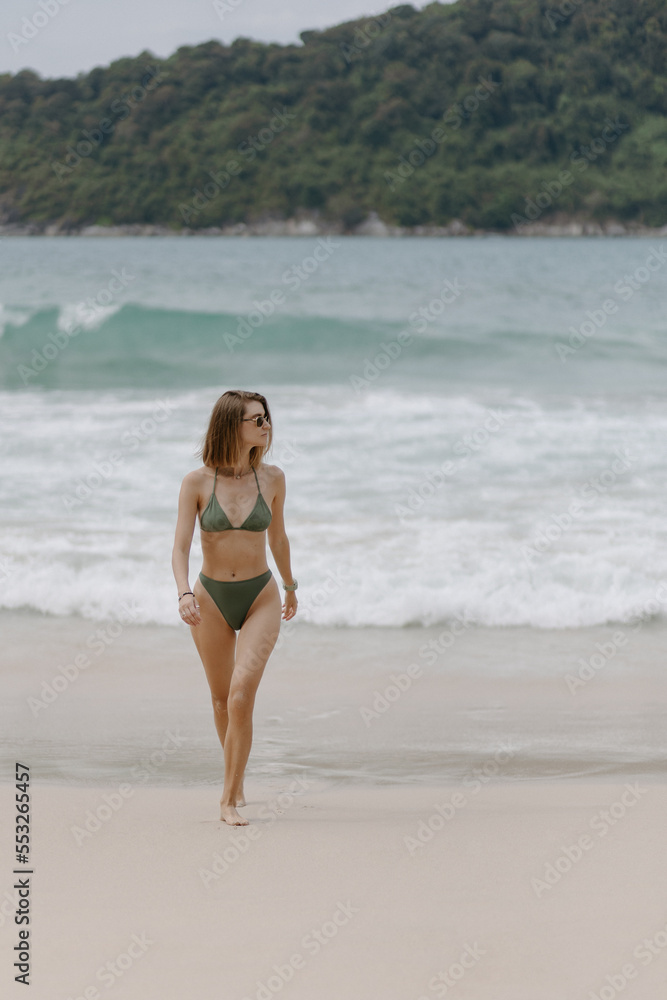 Woman in green bikini walking on beach. Looks to the side. In sunglasses. Against the background of waves. High quality photo