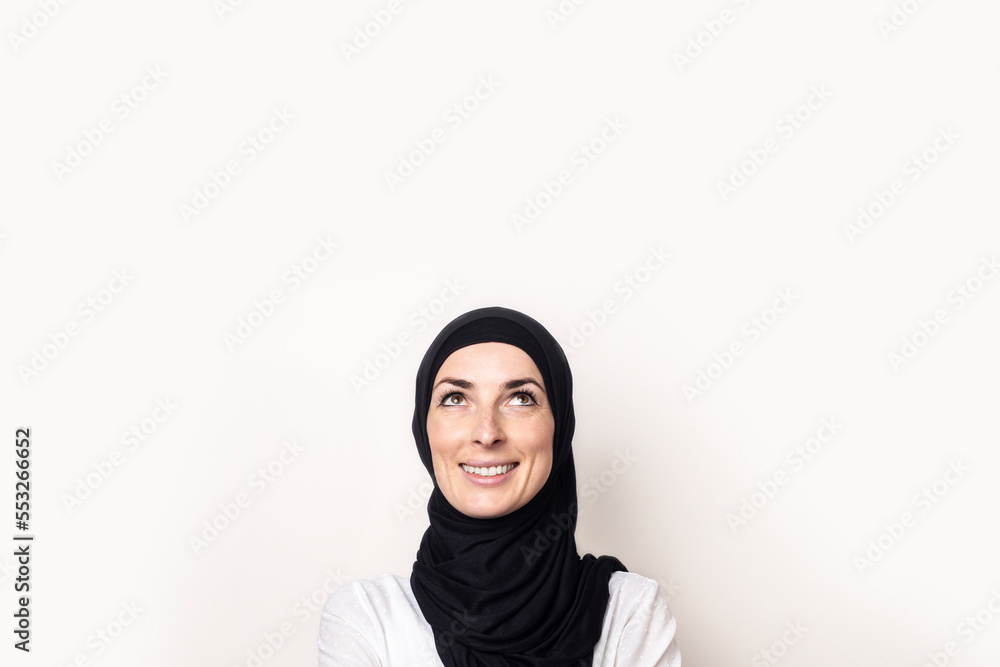 Young Muslim woman dressed in a white shirt and hijab looks up on a light background. Banner Young Muslim woman dressed in a white shirt and hijab looks up on a light background. Banner