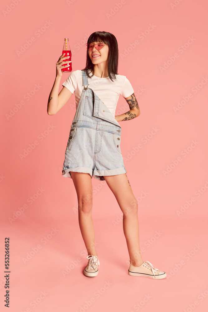 Young stylish girl in trendy sunglasses posing in denim overalls, drinking soda isolated on pink background Young stylish girl in trendy sunglasses posing in denim overalls, drinking soda isolated on pink background