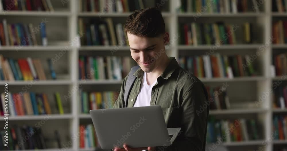 Student guy standing in library holding in hands laptop, typing on keyboard, check online timetable looks focused while make task, finish assignment standing on bookshelves background. Education, tech Student guy standing in library holding in hands laptop, typing on keyboard, check online timetable looks focused while make task, finish assignment standing on bookshelves background. Education, tech