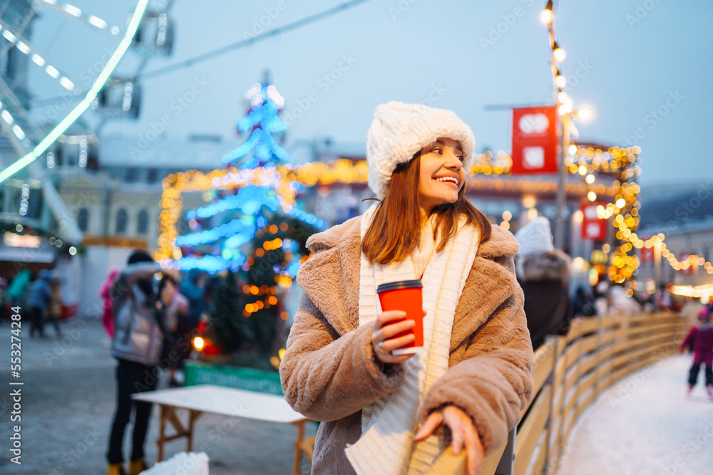 A pretty girl walks around the fair at Christmas, drinks a hot drink from a cup and looks away against the background of beautiful light decorations. A pretty girl walks around the fair at Christmas, drinks a hot drink from a cup and looks away against the background of beautiful light decorations.