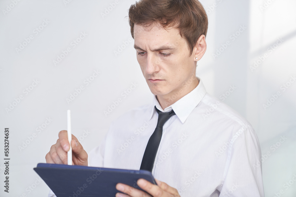 Close-up of a thoughtful young male office worker standing on a light background, with a tablet to search for information. With space to copy. High quality photo Close-up of a thoughtful young male office worker standing on a light background, with a tablet to search for information. With space to copy. High quality photo