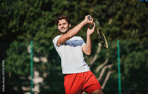 Fototapeta Naklejka Na Ścianę i Meble -  A handsome Caucasian tennis player smiling, carrying his racket and ready to play on an outdoor court, with trees in the background. Concept of joy of playing sports and recreation outdoors.