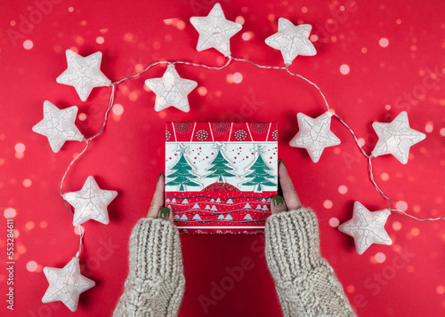 Female hands in white wolen sweater holding present box with green christmas trees on red background with garland shape of star. Festive backdrop for holidays Christmas, New Year. photo