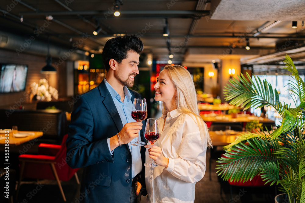 Portrait of cheerful beautiful couple holding glasses of red wine, looking smiling at each other, standing posing in restaurant at evening. Happy young man and woman enjoying nice romantic dinner.