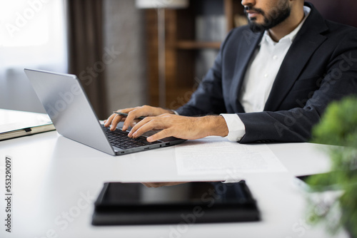 Cropped view of hands of young bearded businessman typing on laptop during work in office. Concentrated adult successful man wearing official suit sitting at wooden desk indoor.