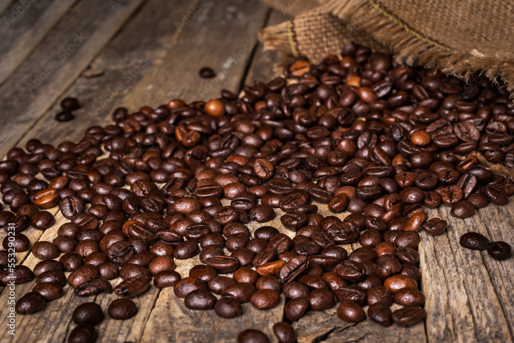 Obraz premium Sack of roasted coffee beans overturned on an old rustic table with the beans scattered on top, close-up.