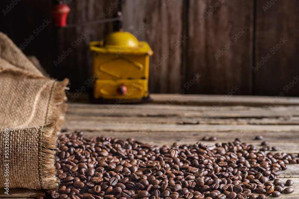 Roasted coffee beans coming out of a sack on a wooden table and in the background an old grinder for grinding coffee.