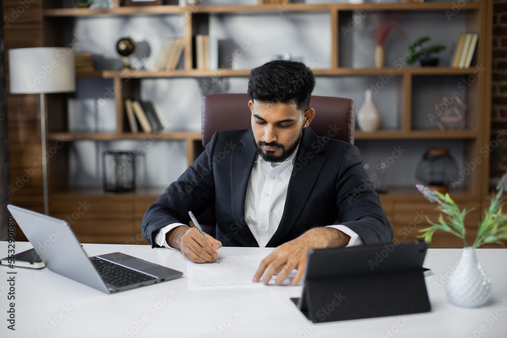 Young bearded male freelancer in business suit signing papers while sitting at table with laptop and tablet in stylish office. Remotely work.