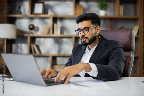 Confident young middle eastern businessman sitting at work table at modern office, speaking with customers, business partners, using laptop during video call, working on marketing research, copy space