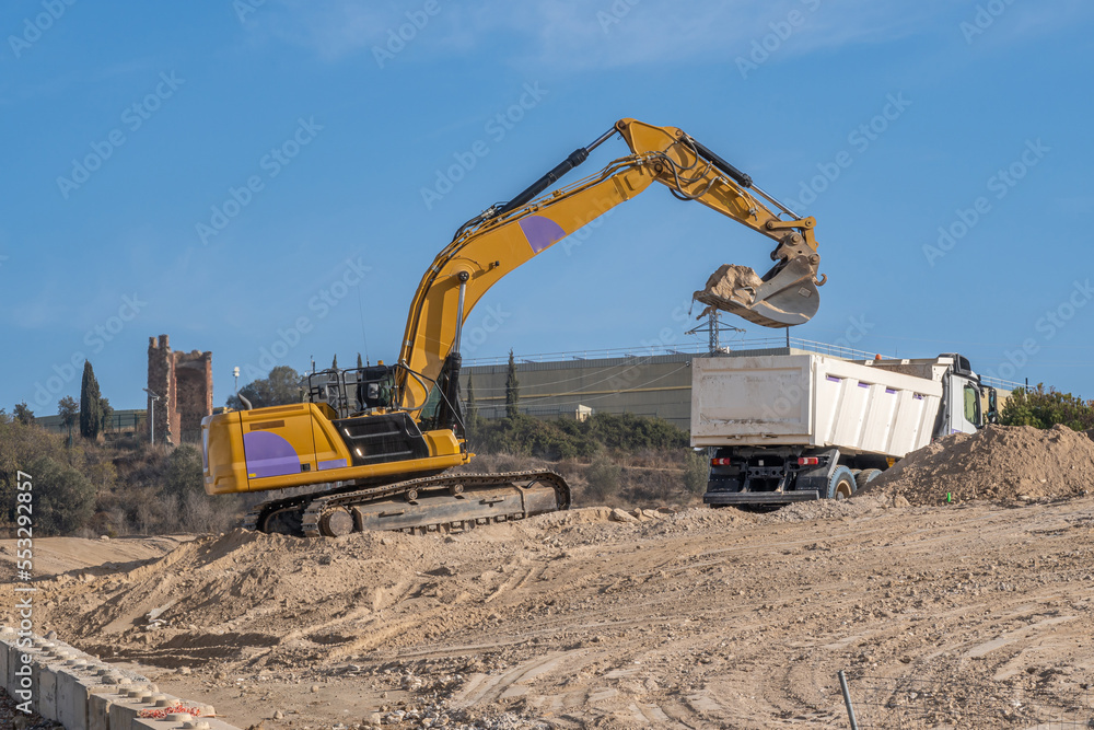 Large yellow excavator depositing sand in a truck bed at a large construction site with disturbed earth Large yellow excavator depositing sand in a truck bed at a large construction site with disturbed earth