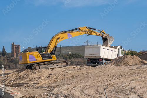 Large yellow excavator depositing sand in a truck bed at a large construction site