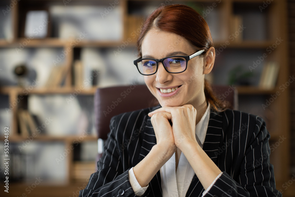 Close-up front view face of cheerful attractive young red hair woman in formal clothing and glasses sitting at office, looking at camera. Pretty redhead caucasian lady remote working or studying.