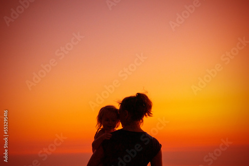 Mom and daughter are standing at sunset. Beautiful picture of motherhood. Silhouettes on a gradient sky