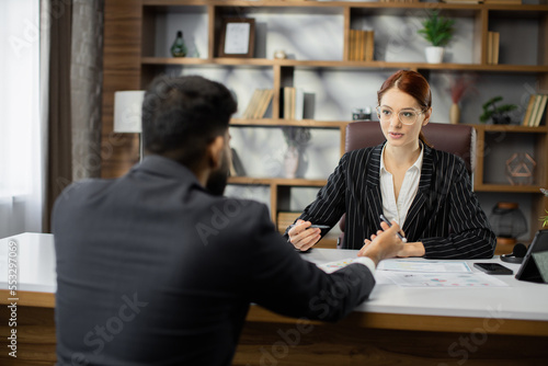 Red hair businesswoman dealer showing papers with new agreement to her handsome bearded businessman customer in a desktop, sitting at table in modern office.