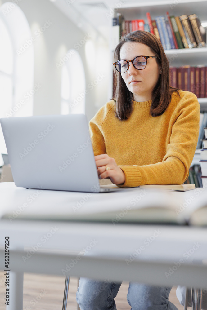thoughtful woman works in library or coworking at laptop and takes notes in notebook. female student is studying online watching educational webinar. bookshelves, place to work thoughtful woman works in library or coworking at laptop and takes notes in notebook. female student is studying online watching educational webinar. bookshelves, place to work