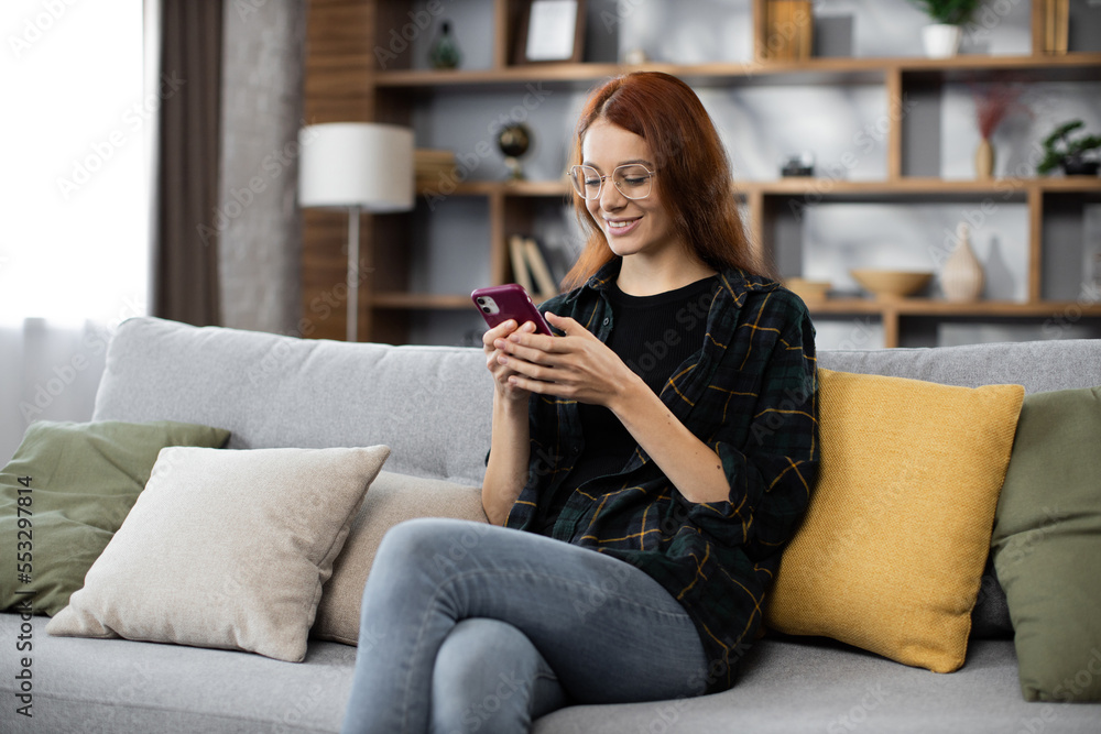 Attractive caucasian red hair woman sitting on couch and using modern smartphone indoors. Female freelancer taking break during working process at home. Attractive caucasian red hair woman sitting on couch and using modern smartphone indoors. Female freelancer taking break during working process at home.