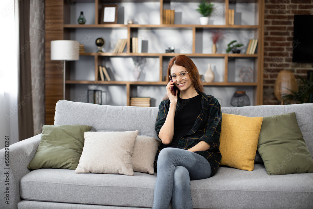 Obraz premium Happy young caucasian woman sitting on cozy sofa with modern smartphone in hands and calling to customers during break. Pretty female resting during working day at home.
