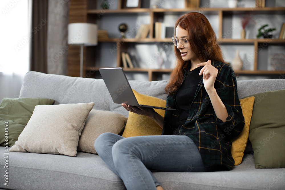 Smiling young woman using laptop, sitting on couch at home, beautiful girl having video call online in social network, having fun, watching movie, freelancer working on computer project Smiling young woman using laptop, sitting on couch at home, beautiful girl having video call online in social network, having fun, watching movie, freelancer working on computer project