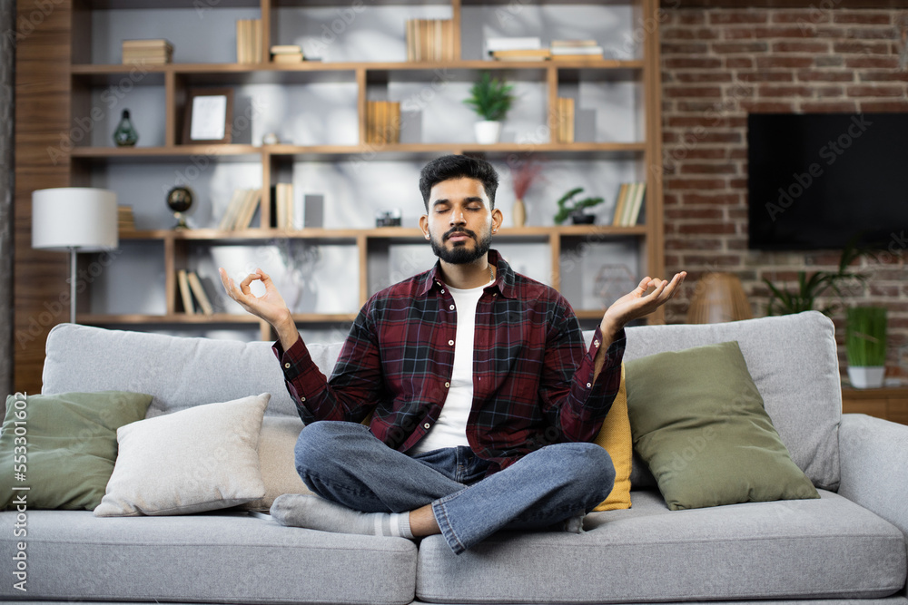 Calm of bearded young man doing yoga lotus pose to meditation and relax on couch during work online at home. Happy male close his eyes after hard work during break and deep breath in yoga posotion