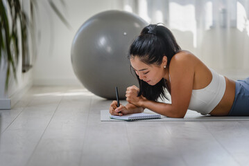 Young Asian yoga woman writing in her notebook. Sitting at home by the window after exercise. Personal growth resolutions 