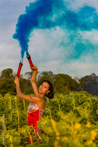 eine junge Frau mit einem rotem Kleid steht in einem Sonnenblumenfeld mit einer blauen Rauchfackel