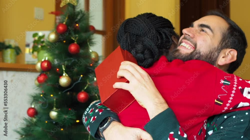 Happy Interracial couple enjoying and hugging in front of a decorated tree wearing christmas sweaters while holding a gift Happy Interracial couple enjoying and hugging in front of a decorated tree wearing christmas sweaters while holding a gift