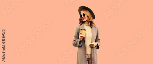Portrait of stylish beautiful young woman model with cup of coffee wearing brown round hat and coat on beige background