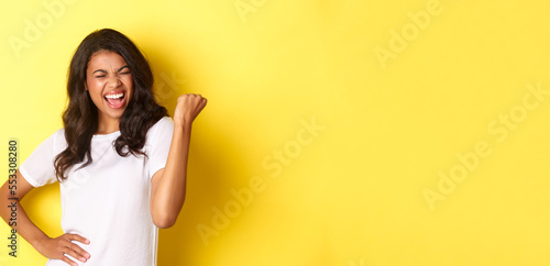 Portrait of happy african-american woman, triumphing and winning, making fist pump and shouting for joy, standing pleased against yellow background