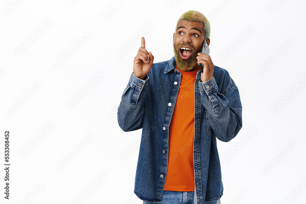 Enthusiastic guy calling someone, pitching an idea, pointing up, standing with mobile phone against white background Enthusiastic guy calling someone, pitching an idea, pointing up, standing with mobile phone against white background